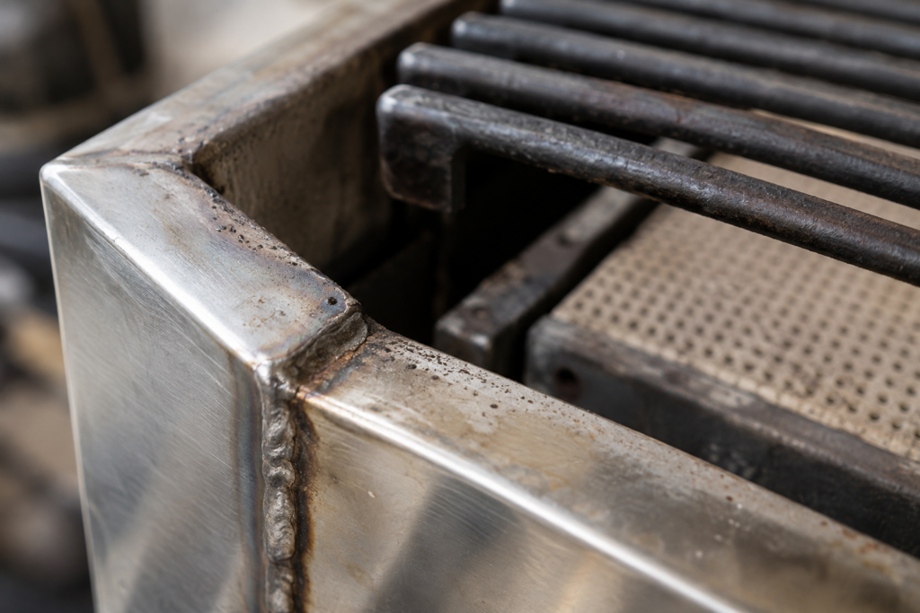 A macro view of the corner of a stainless gas grill firebox, showing the welded seam, a pitted edge, a grate corner and a ceramic radiant.