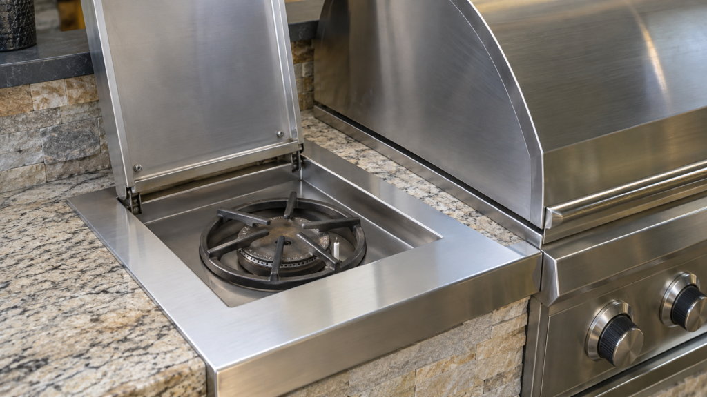 A stainless outdoor grill cart with its small side burner lid raised, the burner ring and igniter electrode visible beneath it.