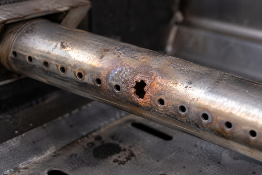 A row of ports along a grill burner tube with one port eaten into a ragged hole and orange rust around it.
