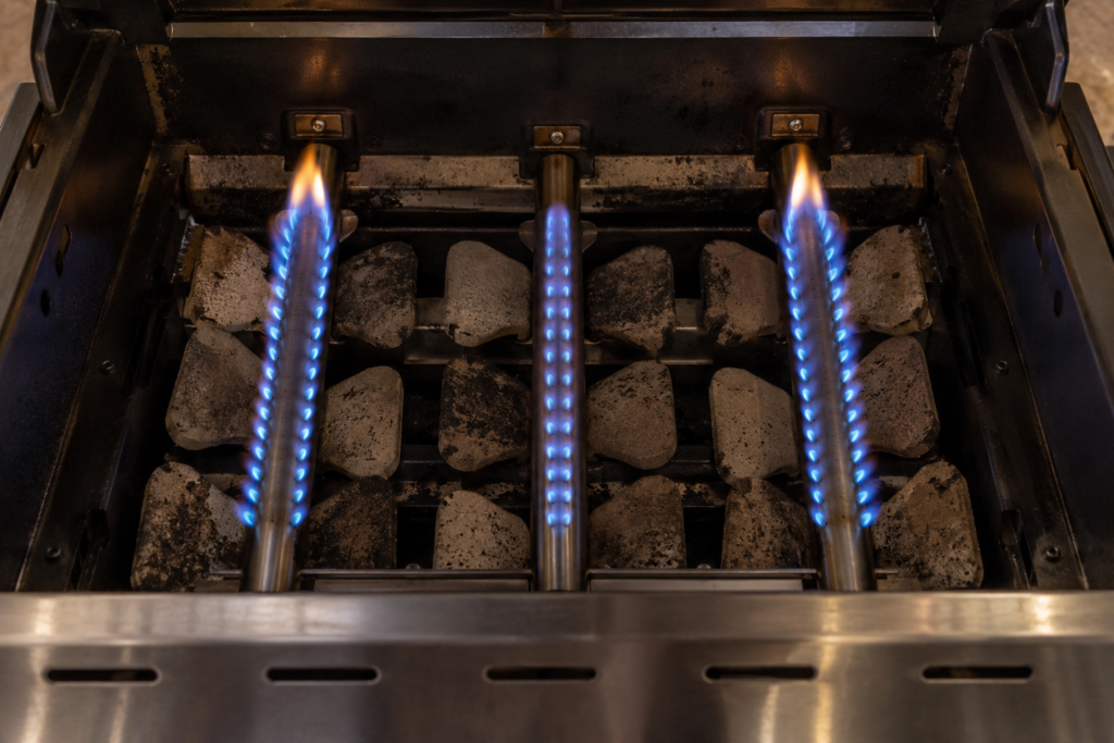 Looking down into the firebox of a gas grill with the cooking grate removed, three burner tubes lit and the middle one showing visibly shorter flames, ceramic briquettes between the tubes, low evening light.